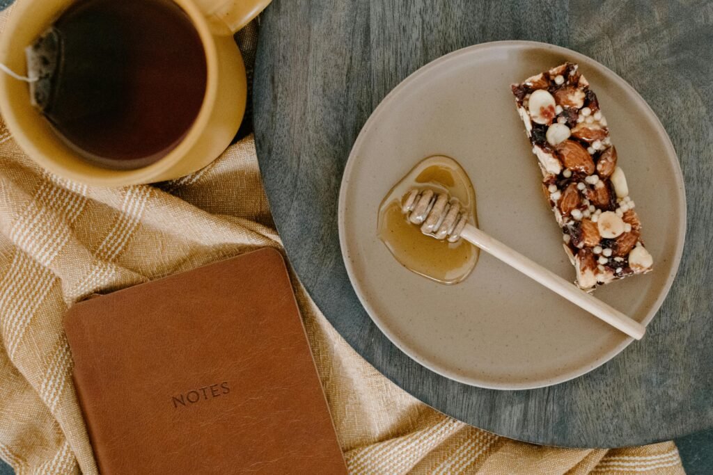 Top view of a nut bar snack, honey dipper, and tea on a wooden table with a notebook.