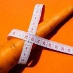 Close-up shot of a carrot wrapped with a measuring tape on a vibrant orange background, symbolizing healthy eating.